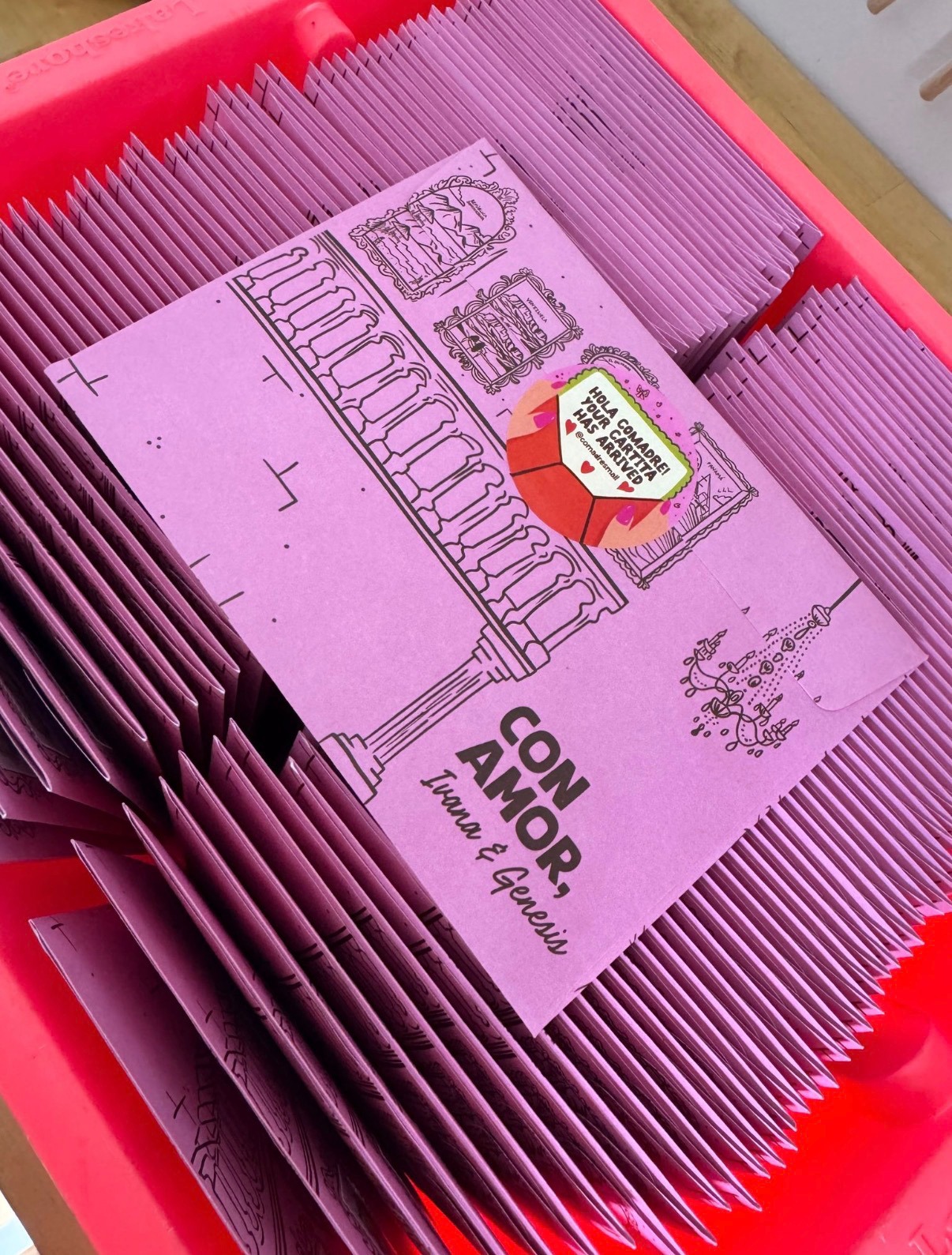 Stacks of pink Las Comadres con amor cartita envelopes in a coral tray